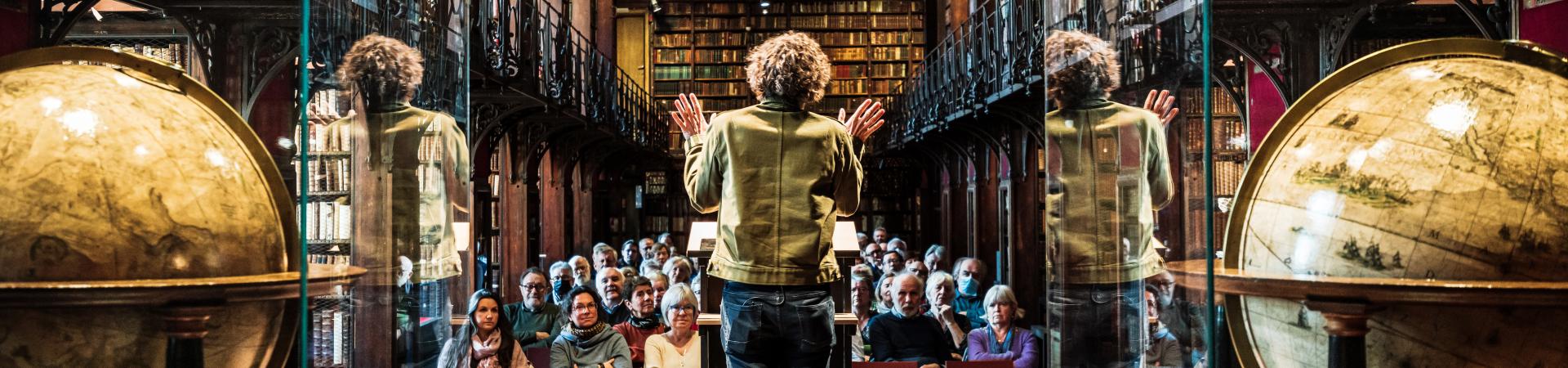 Sfeerbeeld Nottebohmlezing Dirk Draulans aan spreekgestoelte, globes in voorgrond, publiek in achtergrond