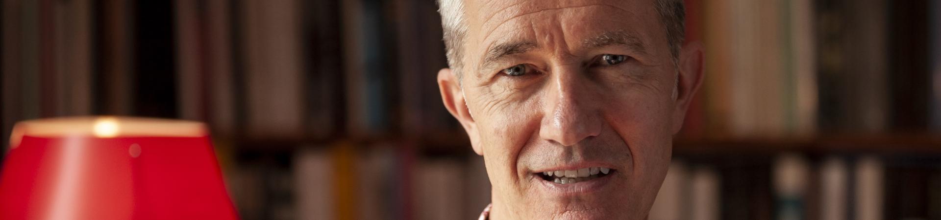 Portretfoto Geoff Dyer naast rode leeslamp en voor boekenkast
