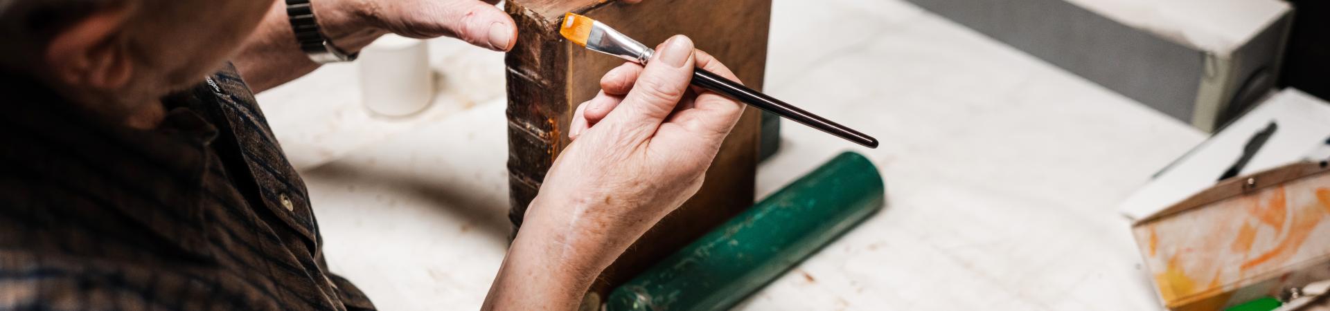 Oudere man herstelt met penseel rug van boek, zittend aan bureau in restauratieatelier.