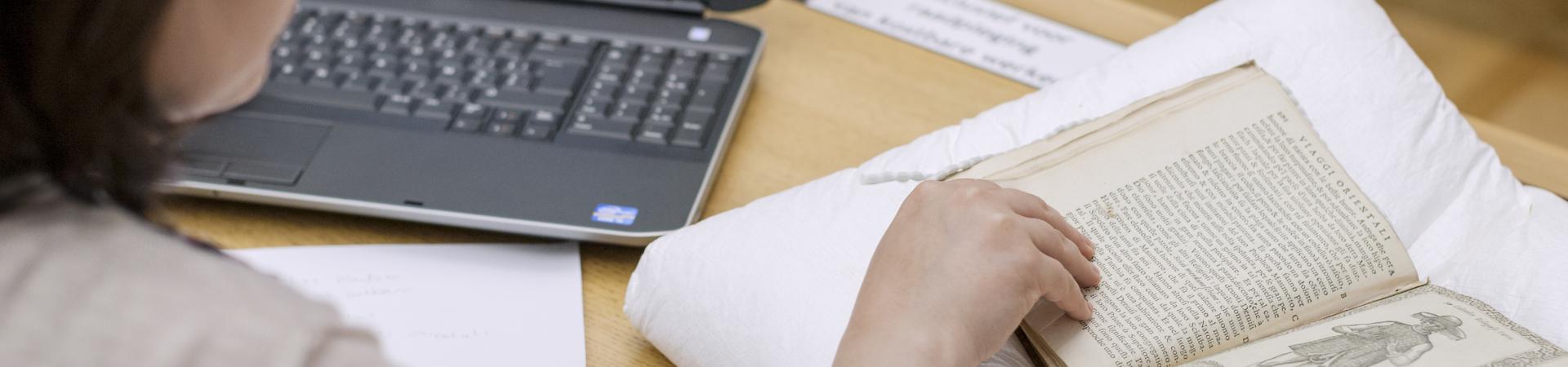 Onderzoekster met laptop en notities bladert door opgevraagde oude druk op leeskussen.