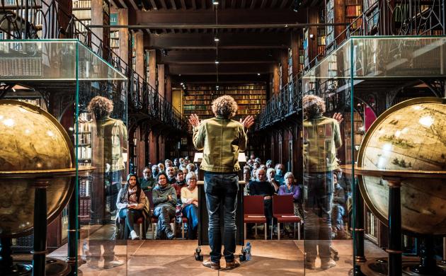 Sfeerbeeld Nottebohmlezing Dirk Draulans aan spreekgestoelte, globes in voorgrond, publiek in achtergrond