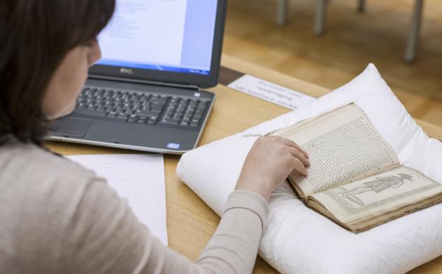 Onderzoekster met laptop en notities bladert door opgevraagde oude druk op leeskussen.