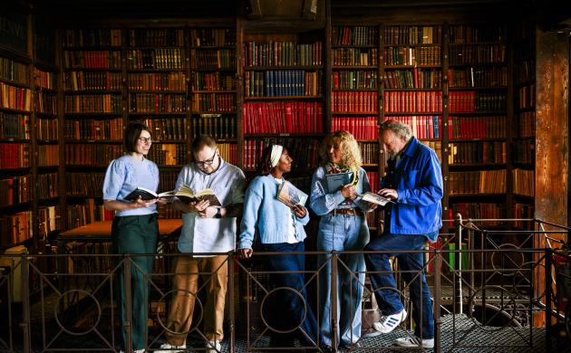 Vijf mensen staan op een ballustrade met op de achtergrond een kast vol oude boeken.